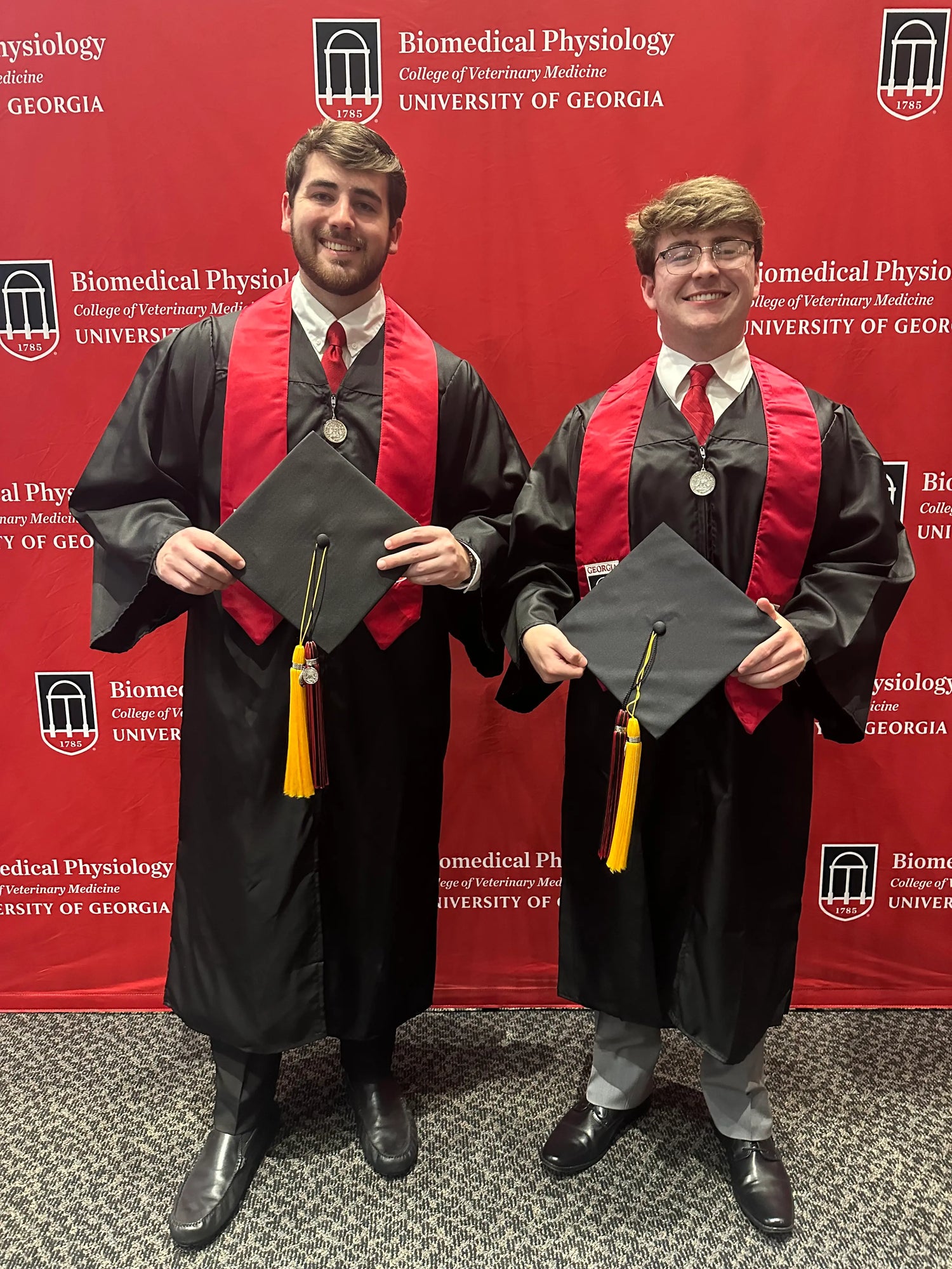 Two graduates (Tyler & Lee) in caps and gowns standing in front of a red backdrop with that says "University of Georgia" "Biomedical Physiology" "College of Veterinary Medicine"